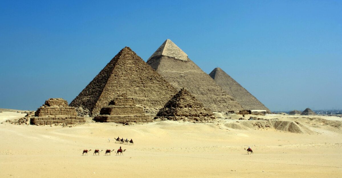 Capture of the iconic Pyramids of Giza under a clear blue sky with camels traversing the sandy desert