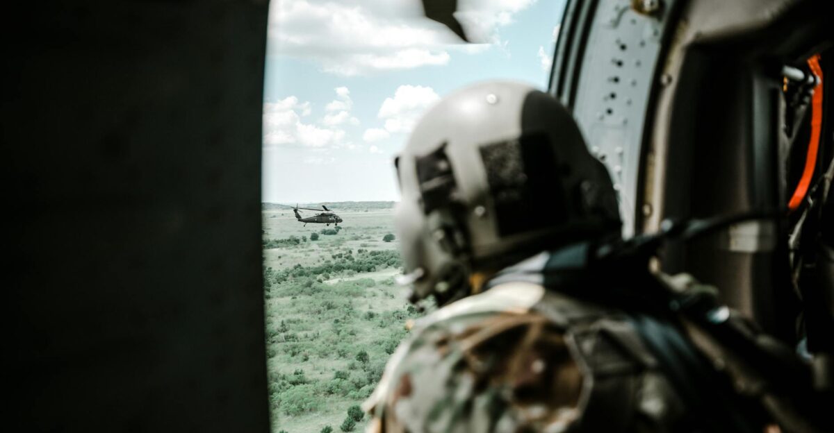 Helmeted soldier in helicopter observing another helicopter over a vast landscape