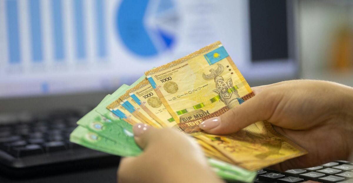 Hands holding Kazakhstan tenge bills with financial chart and graphs on computer screen