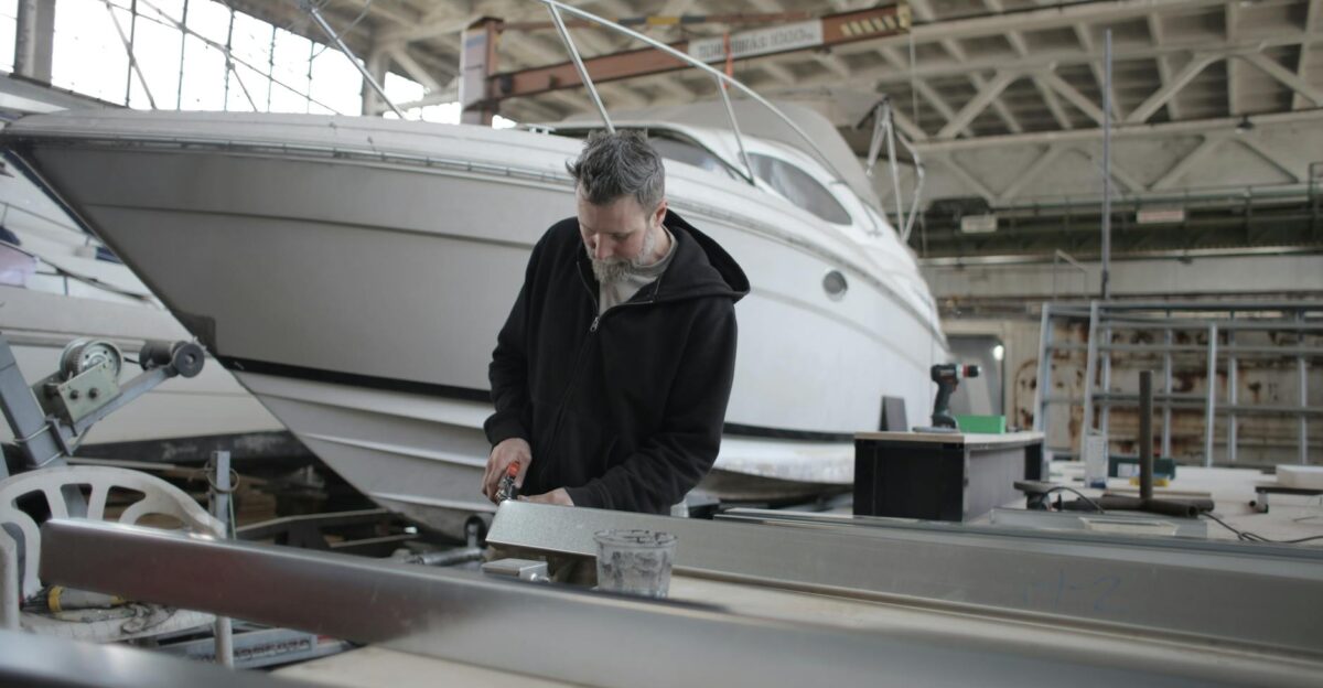 Concentrated bearded adult workman in casual wear working with metal detail near boat in garage