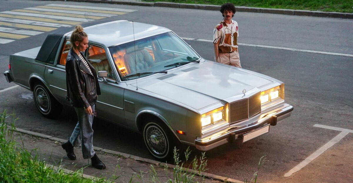 A stylish couple stands by a vintage car parked on a city street in the evening light